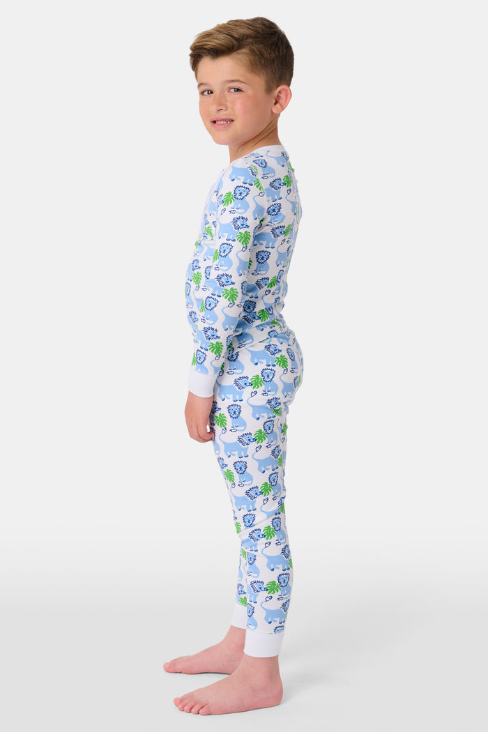 A young child with curly hair stands barefoot, smiling in Roller Rabbit Kids Leo The Lion Pajamas made from soft Pima cotton, against a plain white background.