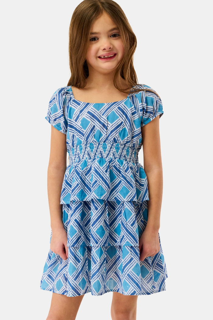 A young girl with long brown hair smiles in a Roller Rabbit Kids Liana Alana Dress, featuring blue and white patterns with a peplum waist, standing against a plain light background.