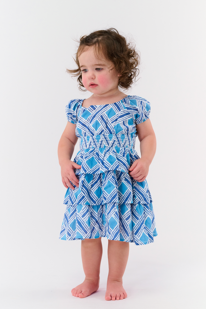A young girl with long brown hair smiles in a Roller Rabbit Kids Liana Alana Dress, featuring blue and white patterns with a peplum waist, standing against a plain light background.