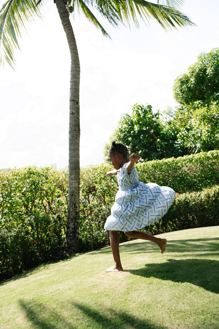 A young girl with long brown hair smiles in a Roller Rabbit Kids Liana Alana Dress, featuring blue and white patterns with a peplum waist, standing against a plain light background.