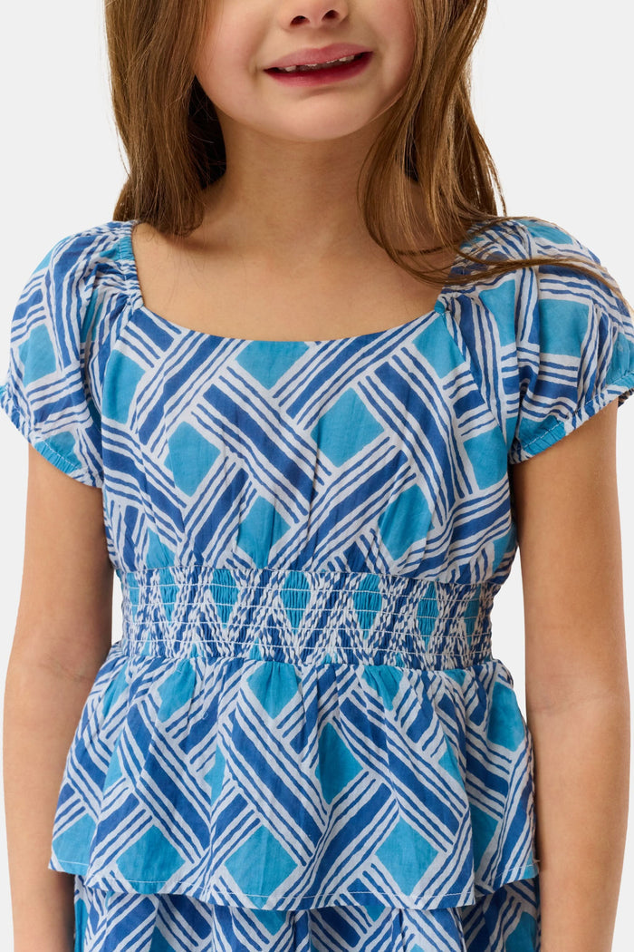 A young girl with long brown hair smiles in a Roller Rabbit Kids Liana Alana Dress, featuring blue and white patterns with a peplum waist, standing against a plain light background.