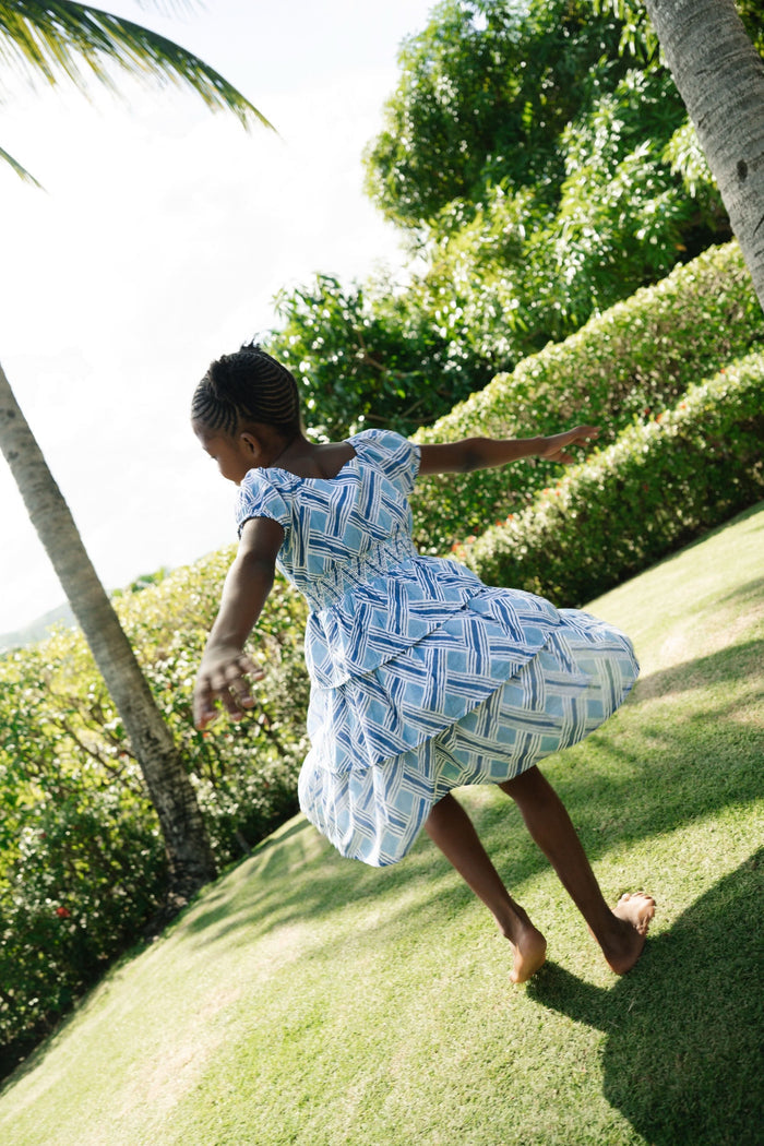 A young girl with long brown hair smiles in a Roller Rabbit Kids Liana Alana Dress, featuring blue and white patterns with a peplum waist, standing against a plain light background.