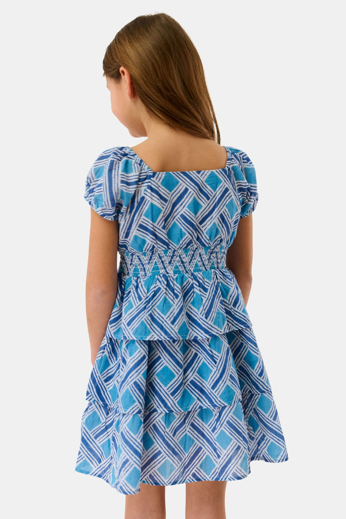A young girl with long brown hair smiles in a Roller Rabbit Kids Liana Alana Dress, featuring blue and white patterns with a peplum waist, standing against a plain light background.
