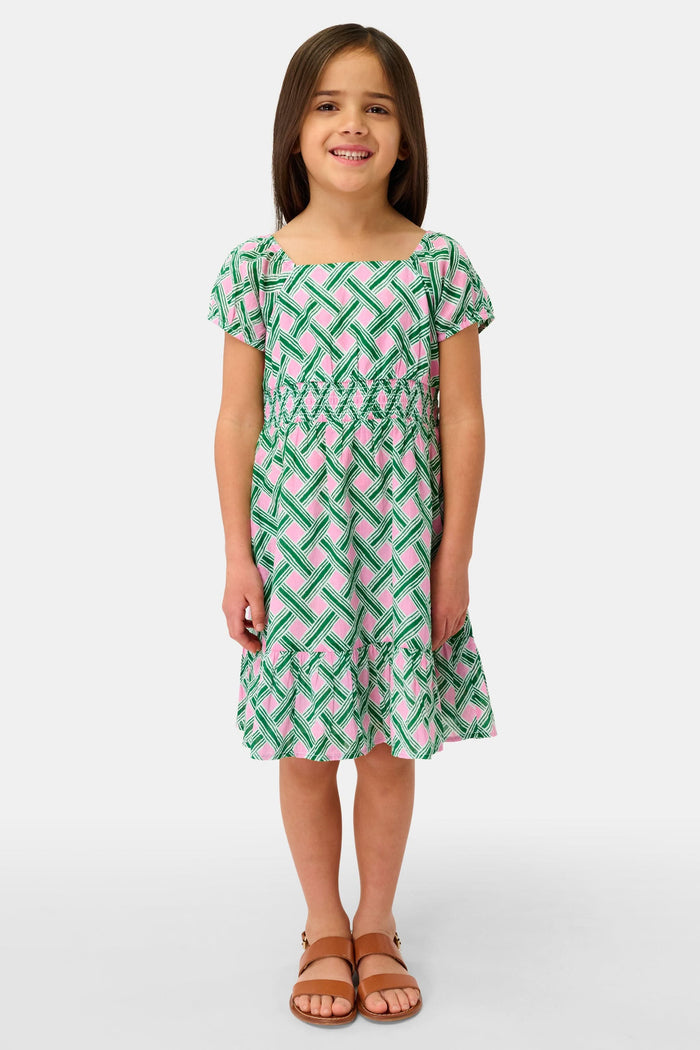 A young girl smiles in the Roller Rabbit Kids Liana Arna Dress, a short-sleeve cotton sundress with a green, pink, and white geometric print and an elastic waist, paired with brown sandals, against a plain white background.
