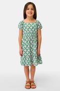 A young girl smiles in the Roller Rabbit Kids Liana Arna Dress, a short-sleeve cotton sundress with a green, pink, and white geometric print and an elastic waist, paired with brown sandals, against a plain white background.