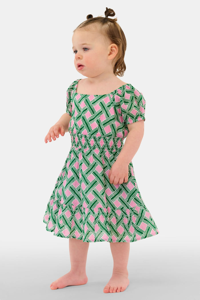 A young girl smiles in the Roller Rabbit Kids Liana Arna Dress, a short-sleeve cotton sundress with a green, pink, and white geometric print and an elastic waist, paired with brown sandals, against a plain white background.
