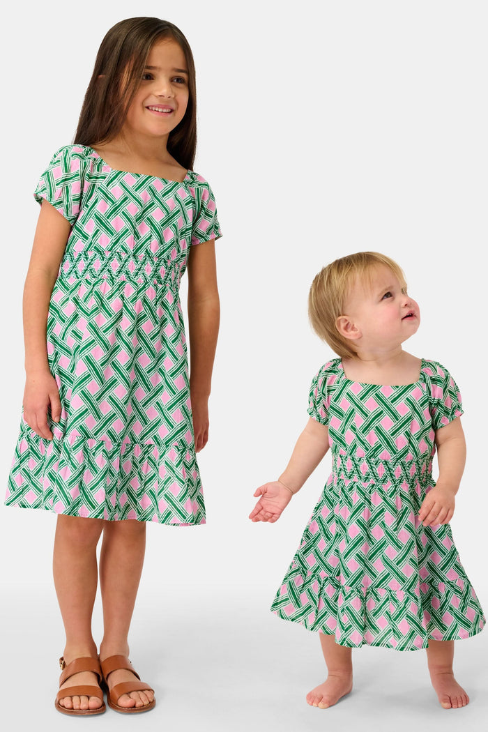 A young girl smiles in the Roller Rabbit Kids Liana Arna Dress, a short-sleeve cotton sundress with a green, pink, and white geometric print and an elastic waist, paired with brown sandals, against a plain white background.