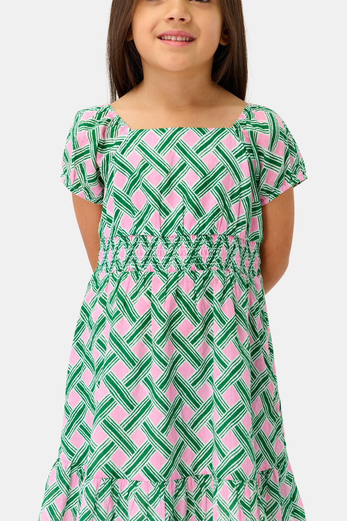 A young girl smiles in the Roller Rabbit Kids Liana Arna Dress, a short-sleeve cotton sundress with a green, pink, and white geometric print and an elastic waist, paired with brown sandals, against a plain white background.