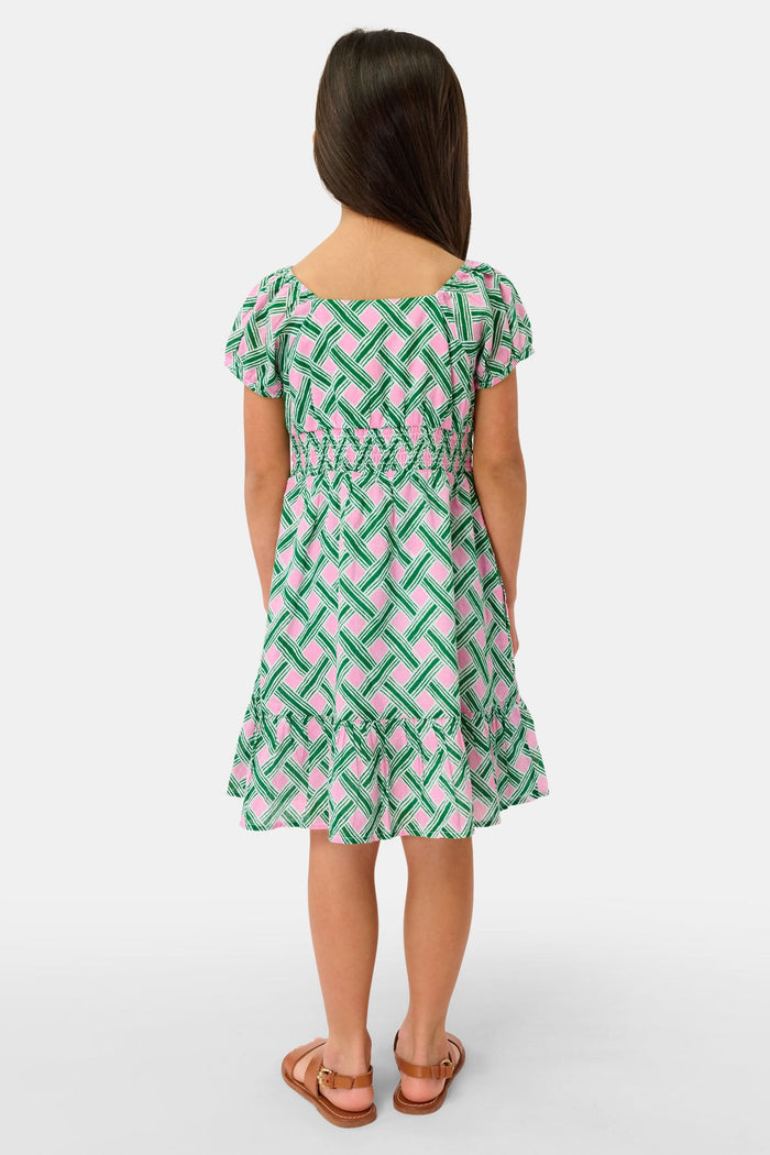 A young girl smiles in the Roller Rabbit Kids Liana Arna Dress, a short-sleeve cotton sundress with a green, pink, and white geometric print and an elastic waist, paired with brown sandals, against a plain white background.