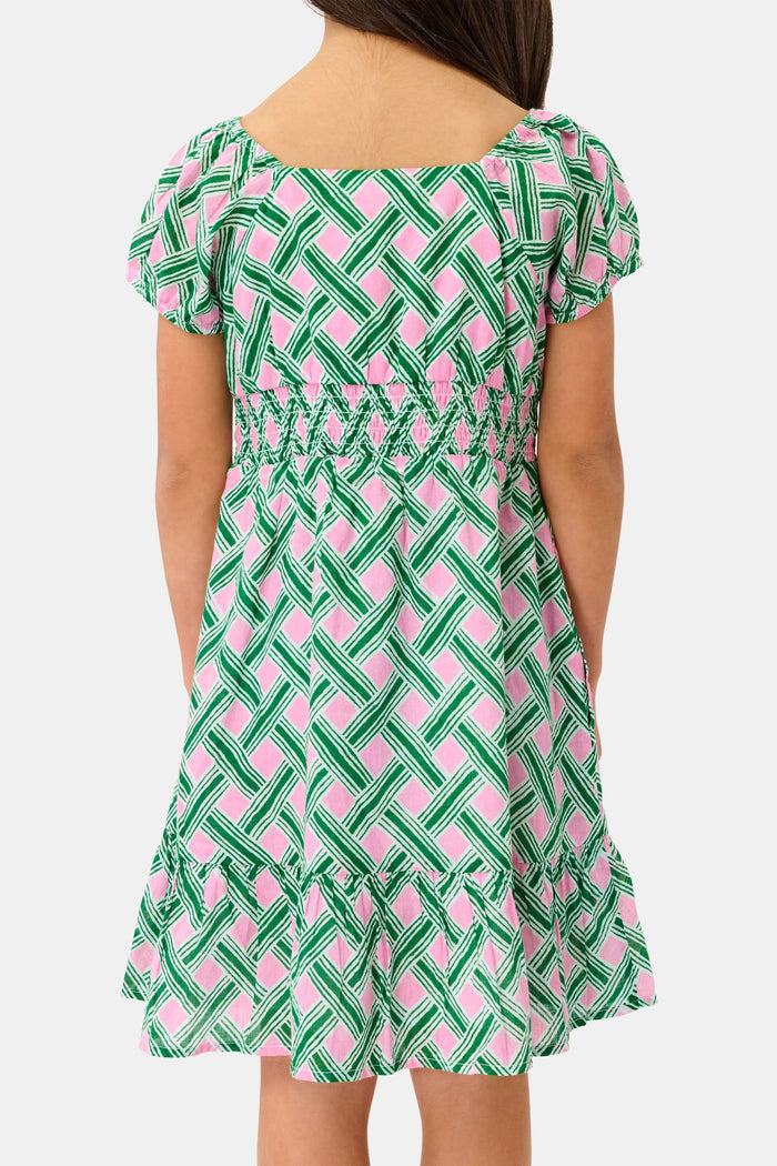 A young girl smiles in the Roller Rabbit Kids Liana Arna Dress, a short-sleeve cotton sundress with a green, pink, and white geometric print and an elastic waist, paired with brown sandals, against a plain white background.