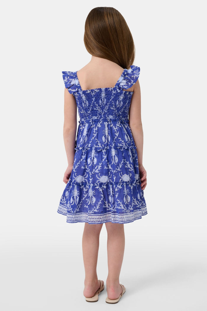 A young girl with straight brown hair smiles while wearing the Roller Rabbit Kids Lobster Trellis Ceri Dress, featuring a blue and white pattern with ruffled sleeves, against a plain white background.