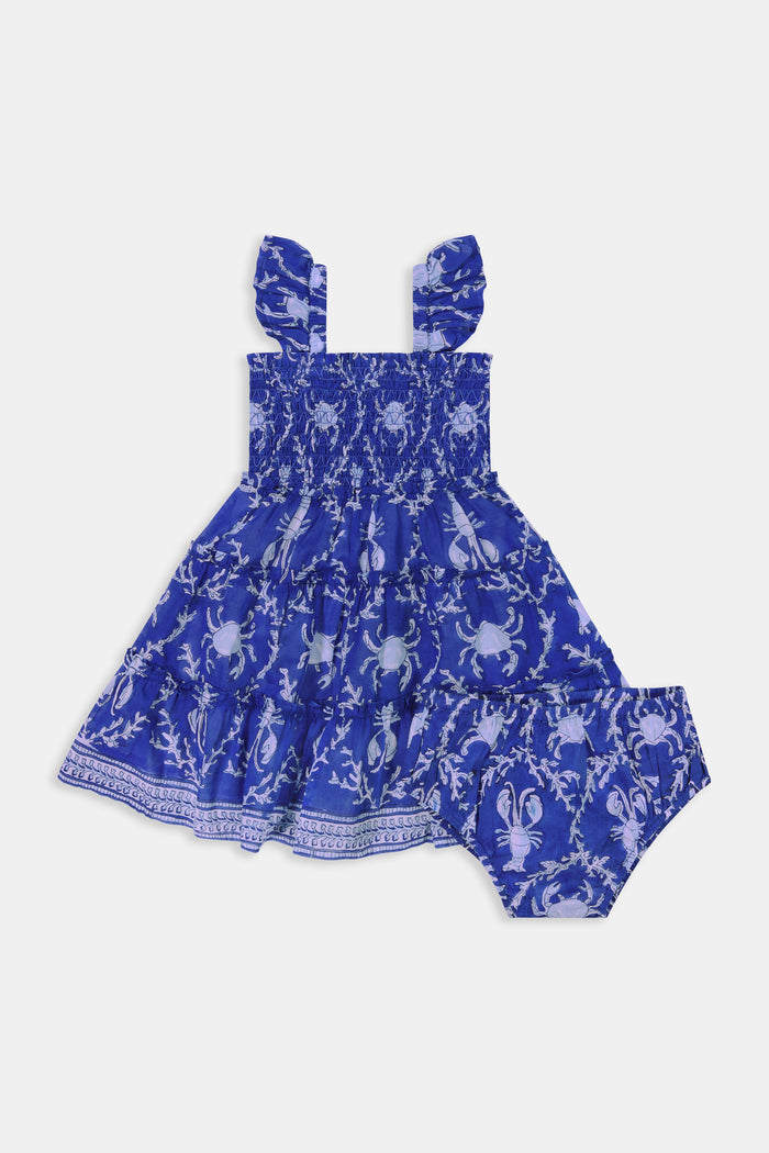 A young girl with straight brown hair smiles while wearing the Roller Rabbit Kids Lobster Trellis Ceri Dress, featuring a blue and white pattern with ruffled sleeves, against a plain white background.