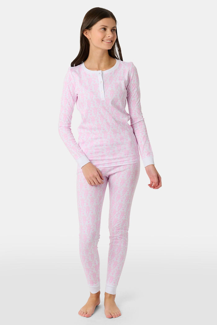 A woman stands barefoot against a plain light background, smiling and looking aside while wearing Roller Rabbit Kids Lovely Monkey Pajamas—long-sleeve, button-collar light pink Pima cotton with a monkey print.