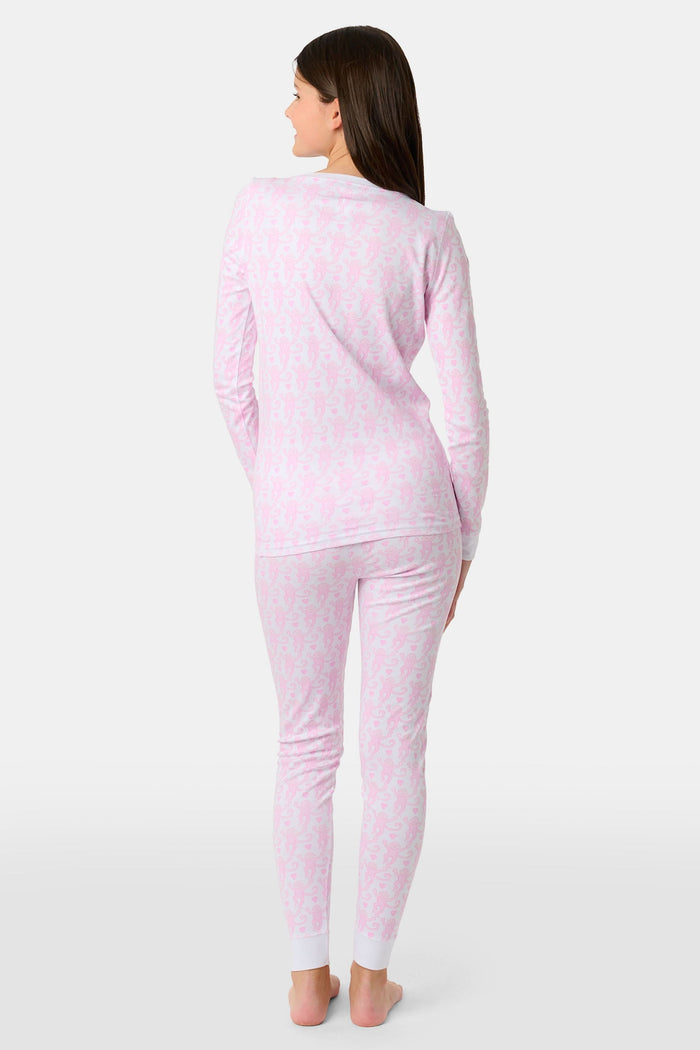 A woman stands barefoot against a plain light background, smiling and looking aside while wearing Roller Rabbit Kids Lovely Monkey Pajamas—long-sleeve, button-collar light pink Pima cotton with a monkey print.