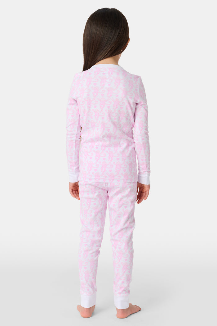 A woman stands barefoot against a plain light background, smiling and looking aside while wearing Roller Rabbit Kids Lovely Monkey Pajamas—long-sleeve, button-collar light pink Pima cotton with a monkey print.