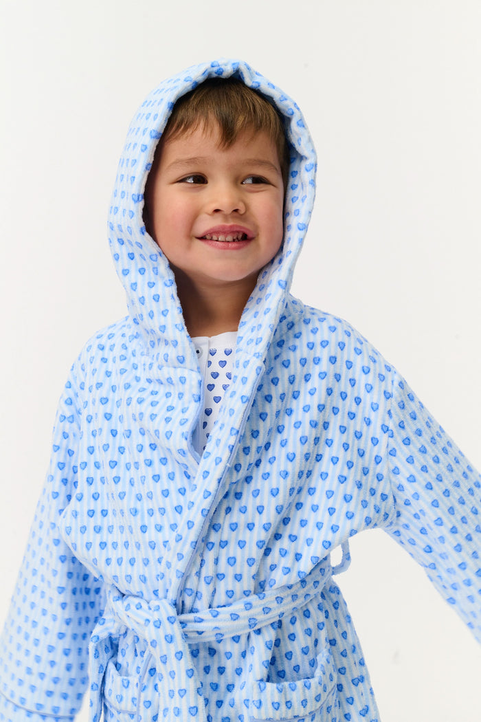 A young child smiles while standing on one leg, wearing the Roller Rabbit Kids Lovely Stripe Max Bathrobe with matching pajamas, in light blue and white, against a plain white background.
