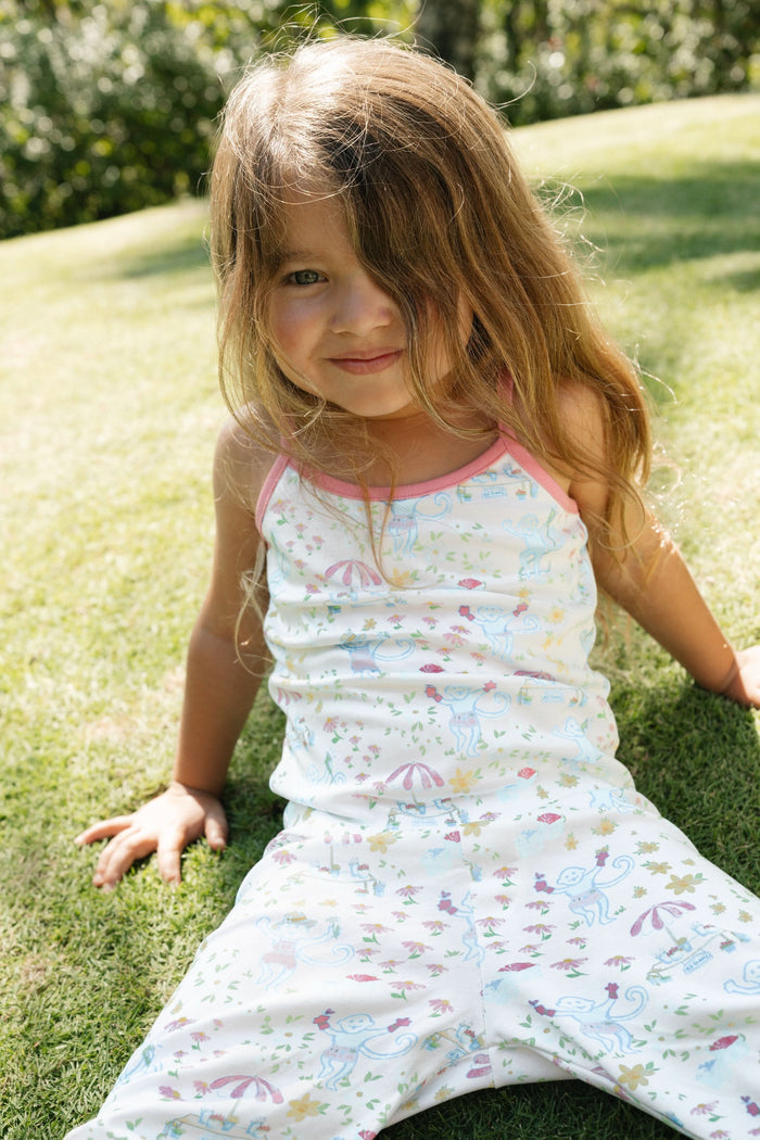 A young girl smiles barefoot against a white background, wearing the Roller Rabbit Kids May Flowers Noa Set—light Pima Cotton pajamas trimmed in pink with a playful floral print.