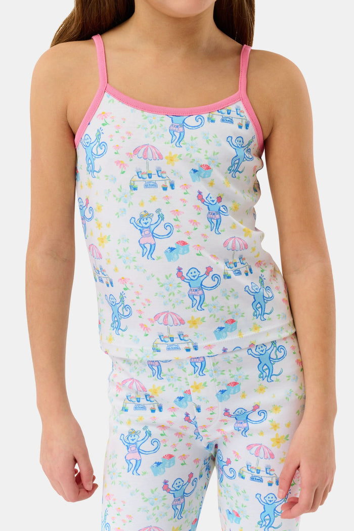 A young girl smiles barefoot against a white background, wearing the Roller Rabbit Kids May Flowers Noa Set—light Pima Cotton pajamas trimmed in pink with a playful floral print.
