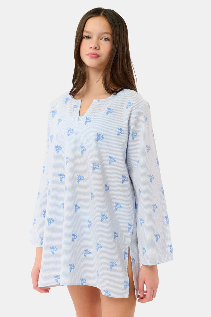 A young girl with long brown hair smiles while wearing a Roller Rabbit Kids Mini Monkey Kurta, made of cotton seersucker with a light blue and white striped pattern, blue floral accents, and long sleeves, against a plain white background.