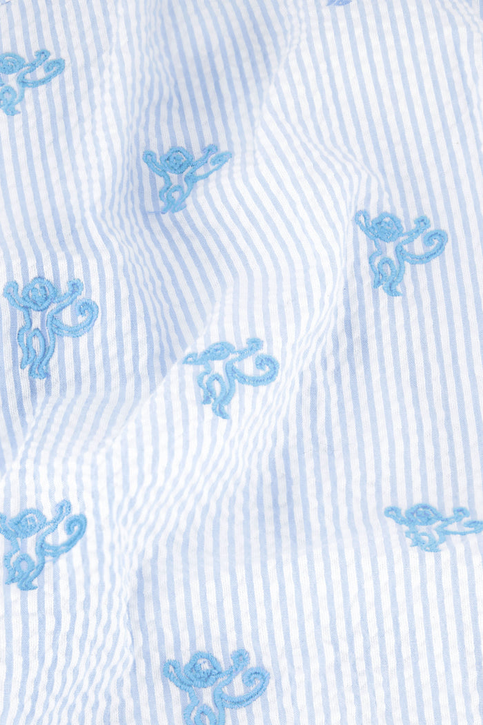 A woman poses barefoot against a white background in the Roller Rabbit Mini Monkey Liza Set—a matching short-sleeve, collared pajama set featuring white fabric with a light blue floral print, blue piping, and monkey embroidery.