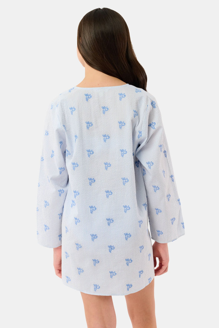 A young girl with long brown hair smiles while wearing a Roller Rabbit Kids Mini Monkey Kurta, made of cotton seersucker with a light blue and white striped pattern, blue floral accents, and long sleeves, against a plain white background.