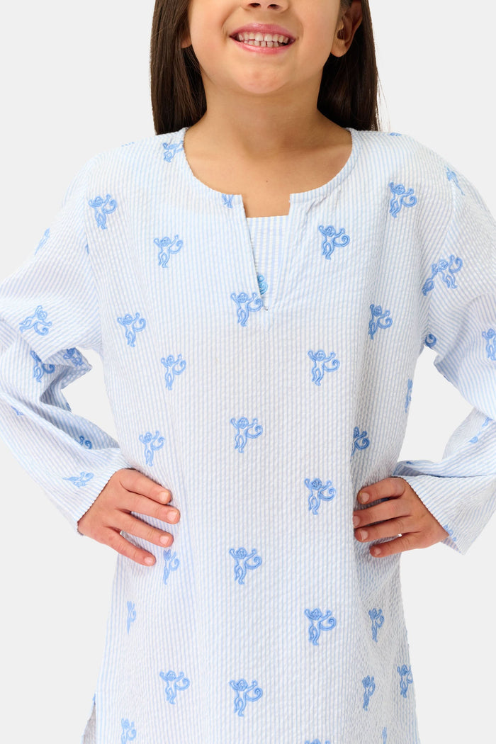 A young girl with long brown hair smiles while wearing a Roller Rabbit Kids Mini Monkey Kurta, made of cotton seersucker with a light blue and white striped pattern, blue floral accents, and long sleeves, against a plain white background.