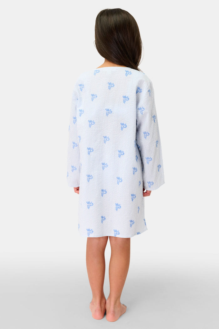 A young girl with long brown hair smiles while wearing a Roller Rabbit Kids Mini Monkey Kurta, made of cotton seersucker with a light blue and white striped pattern, blue floral accents, and long sleeves, against a plain white background.