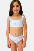 A young girl with long brown hair is smiling and standing, wearing the Roller Rabbit Kids Mini Monkey Rory Bikini—a blue and white striped seersucker bikini with ruffled straps and small blue floral patterns on a plain background.