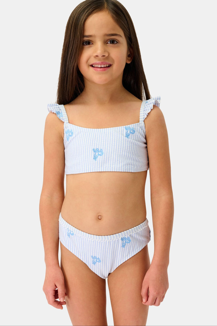 A young girl with long brown hair is smiling and standing, wearing the Roller Rabbit Kids Mini Monkey Rory Bikini—a blue and white striped seersucker bikini with ruffled straps and small blue floral patterns on a plain background.