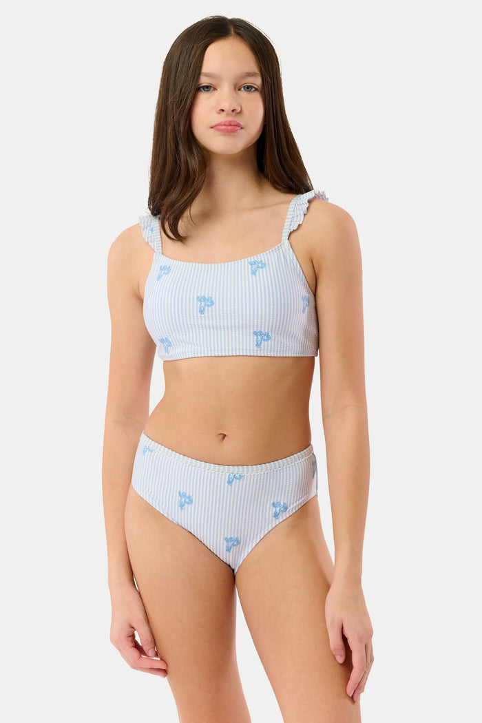 A young girl with long brown hair is smiling and standing, wearing the Roller Rabbit Kids Mini Monkey Rory Bikini—a blue and white striped seersucker bikini with ruffled straps and small blue floral patterns on a plain background.