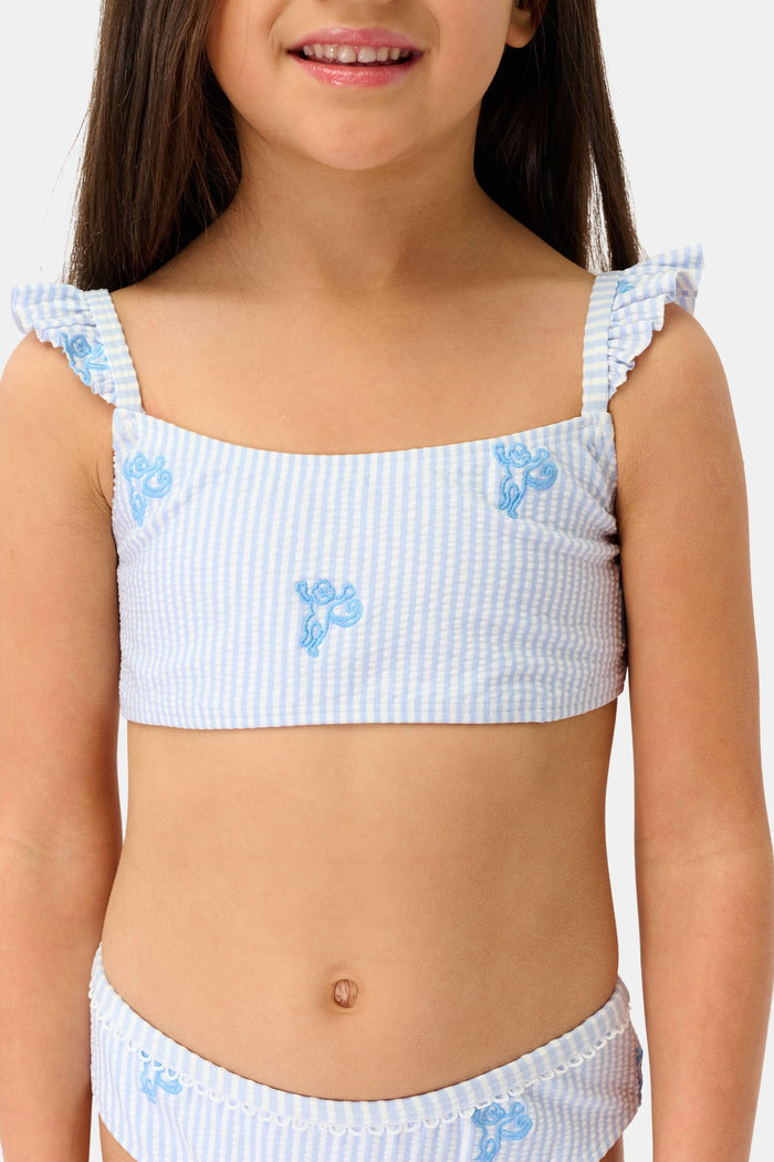 A young girl with long brown hair is smiling and standing, wearing the Roller Rabbit Kids Mini Monkey Rory Bikini—a blue and white striped seersucker bikini with ruffled straps and small blue floral patterns on a plain background.