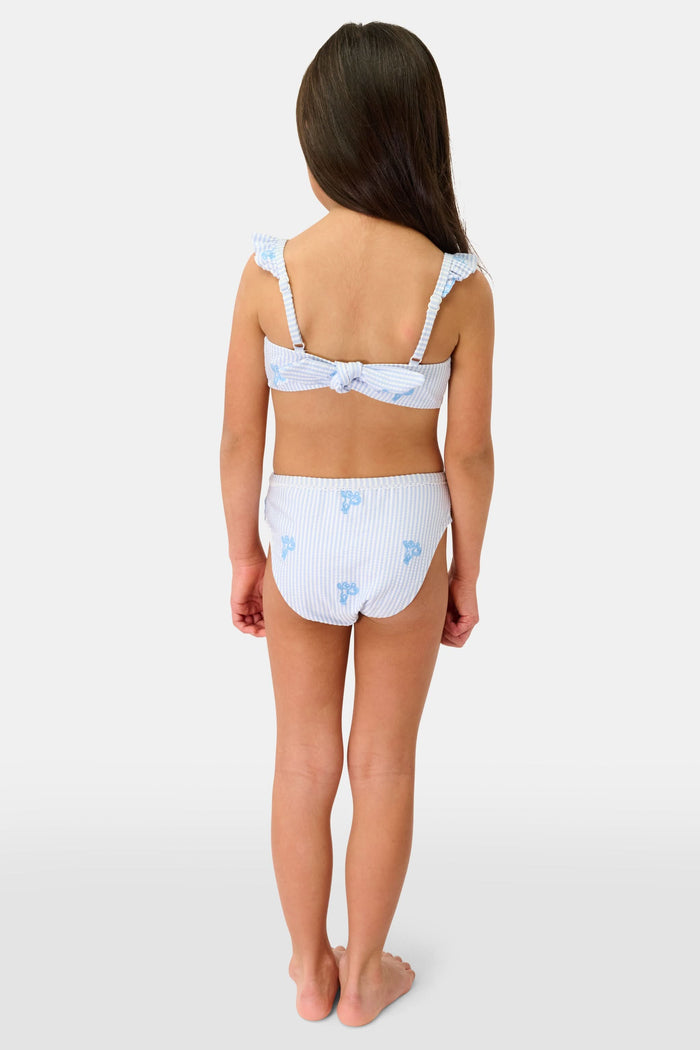 A young girl with long brown hair is smiling and standing, wearing the Roller Rabbit Kids Mini Monkey Rory Bikini—a blue and white striped seersucker bikini with ruffled straps and small blue floral patterns on a plain background.