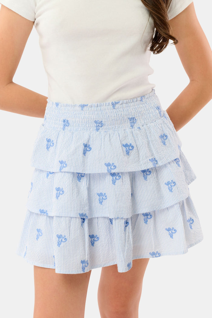 A person is shown from shoulders to upper legs wearing a white short-sleeve top and the Roller Rabbit Kids Mini Monkey Sami Skirt, a baby blue seersucker skirt with small embroidered monkeys.