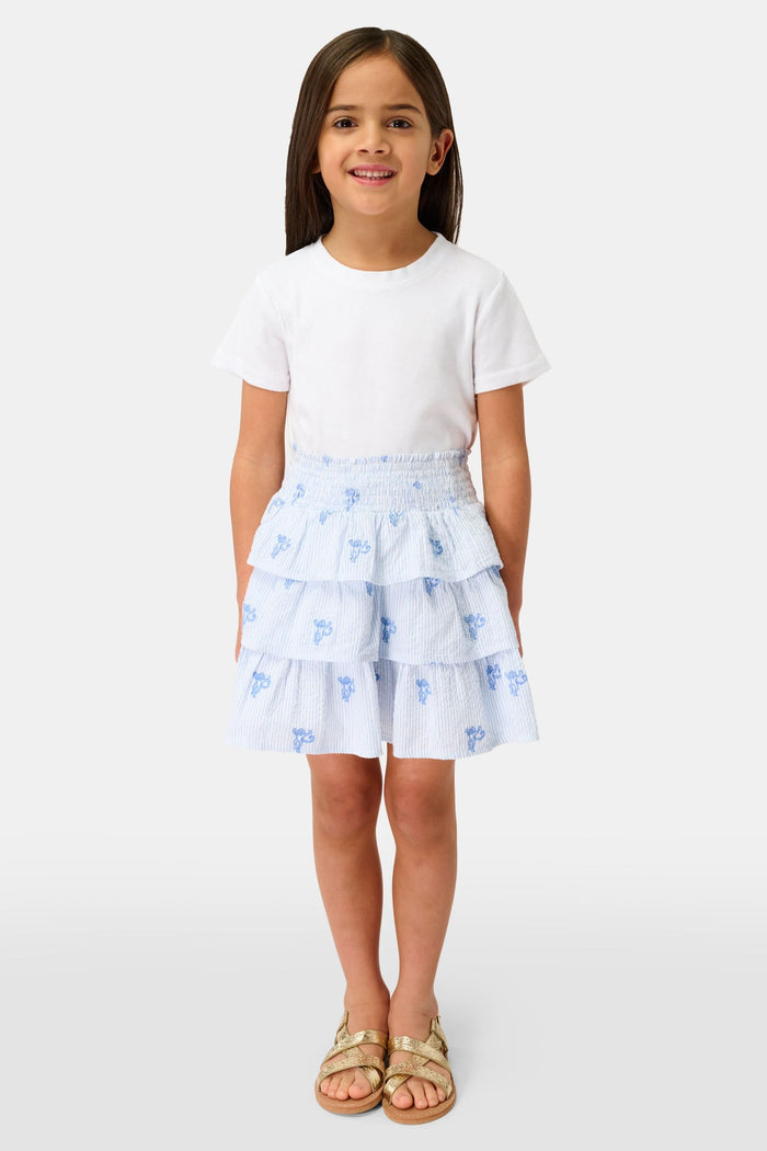 A person is shown from shoulders to upper legs wearing a white short-sleeve top and the Roller Rabbit Kids Mini Monkey Sami Skirt, a baby blue seersucker skirt with small embroidered monkeys.