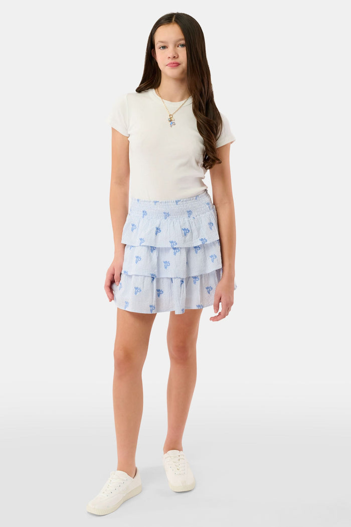 A person is shown from shoulders to upper legs wearing a white short-sleeve top and the Roller Rabbit Kids Mini Monkey Sami Skirt, a baby blue seersucker skirt with small embroidered monkeys.