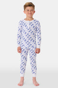 A young boy stands barefoot and smiling in Roller Rabbit's Kids Moby Pajamas—white Pima cotton pajamas with blue car prints, featuring long sleeves and pants—against a plain white background.