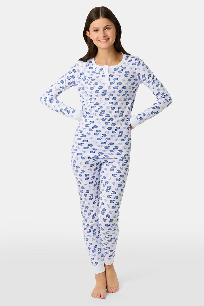 A young boy stands barefoot and smiling in Roller Rabbit's Kids Moby Pajamas—white Pima cotton pajamas with blue car prints, featuring long sleeves and pants—against a plain white background.