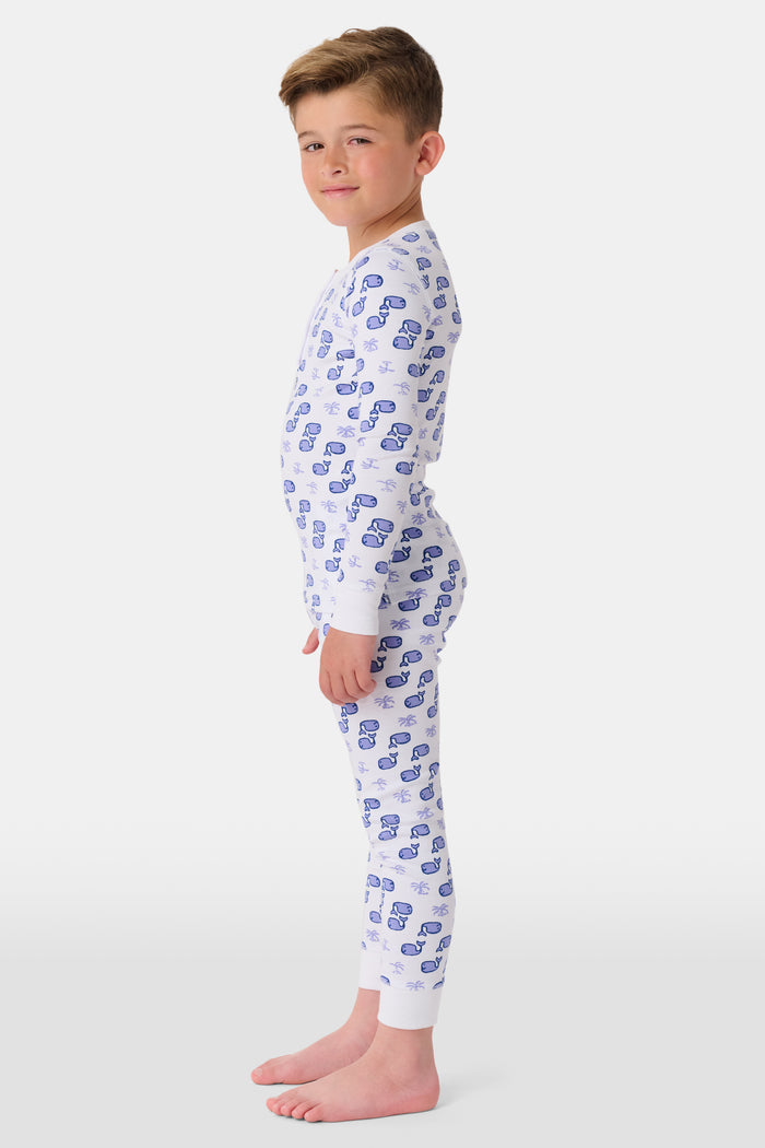 A young boy stands barefoot and smiling in Roller Rabbit's Kids Moby Pajamas—white Pima cotton pajamas with blue car prints, featuring long sleeves and pants—against a plain white background.