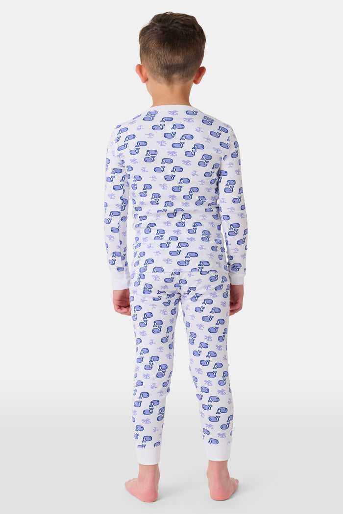 A young boy stands barefoot and smiling in Roller Rabbit's Kids Moby Pajamas—white Pima cotton pajamas with blue car prints, featuring long sleeves and pants—against a plain white background.