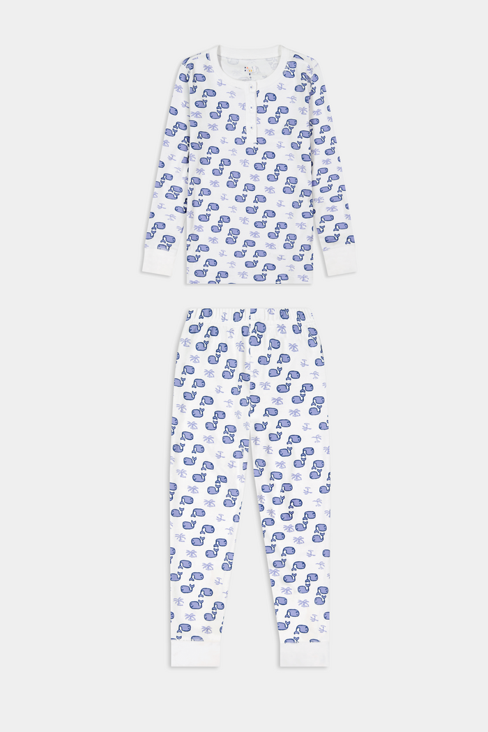 A young boy stands barefoot and smiling in Roller Rabbit's Kids Moby Pajamas—white Pima cotton pajamas with blue car prints, featuring long sleeves and pants—against a plain white background.