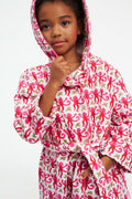 A young girl models the Roller Rabbit Kids Monkey Cosmo Bathrobe, featuring a pink and red monkey print with a hood. She holds the collar with one hand and has her other hand in her pocket, standing against a plain white background.