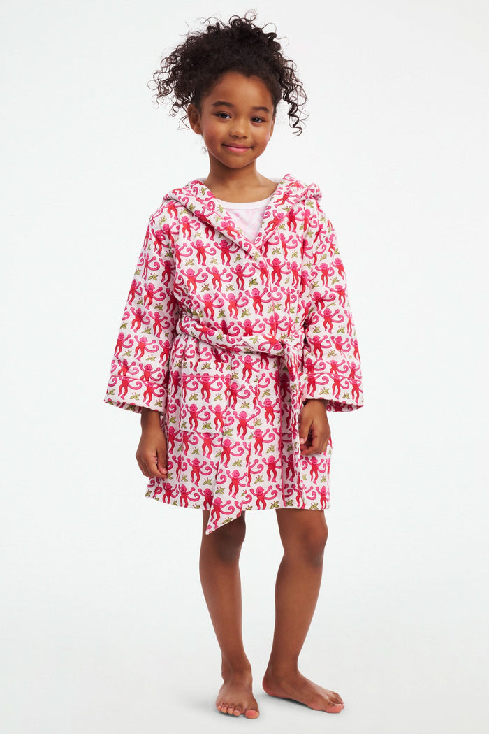 A young girl models the Roller Rabbit Kids Monkey Cosmo Bathrobe, featuring a pink and red monkey print with a hood. She holds the collar with one hand and has her other hand in her pocket, standing against a plain white background.