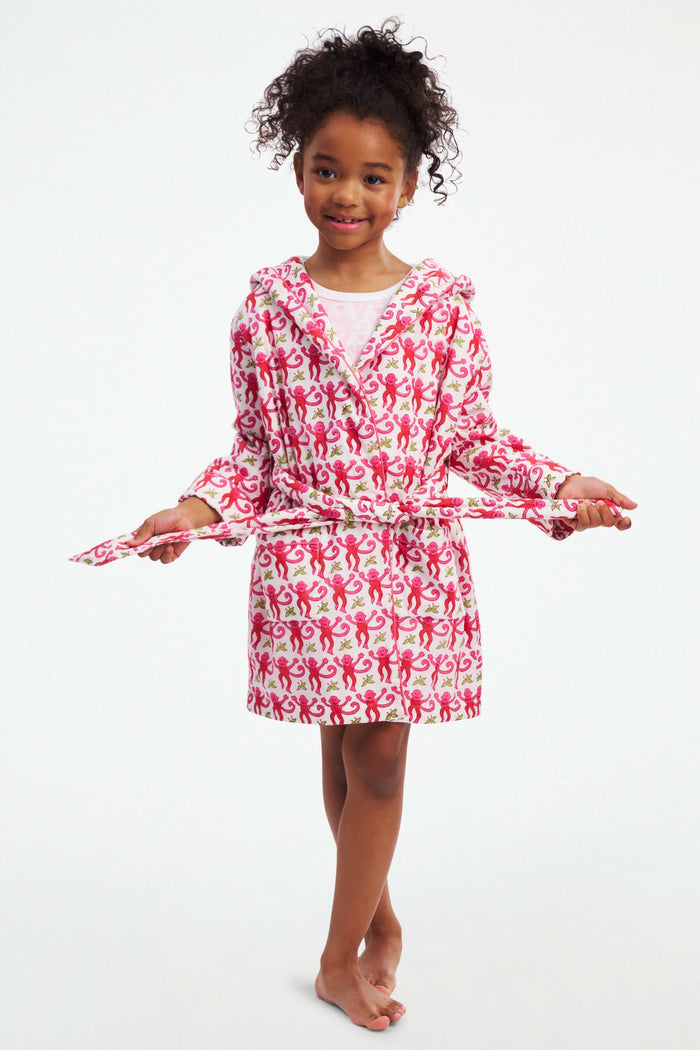 A young girl models the Roller Rabbit Kids Monkey Cosmo Bathrobe, featuring a pink and red monkey print with a hood. She holds the collar with one hand and has her other hand in her pocket, standing against a plain white background.