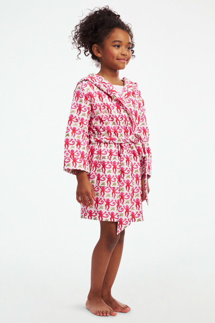 A young girl models the Roller Rabbit Kids Monkey Cosmo Bathrobe, featuring a pink and red monkey print with a hood. She holds the collar with one hand and has her other hand in her pocket, standing against a plain white background.