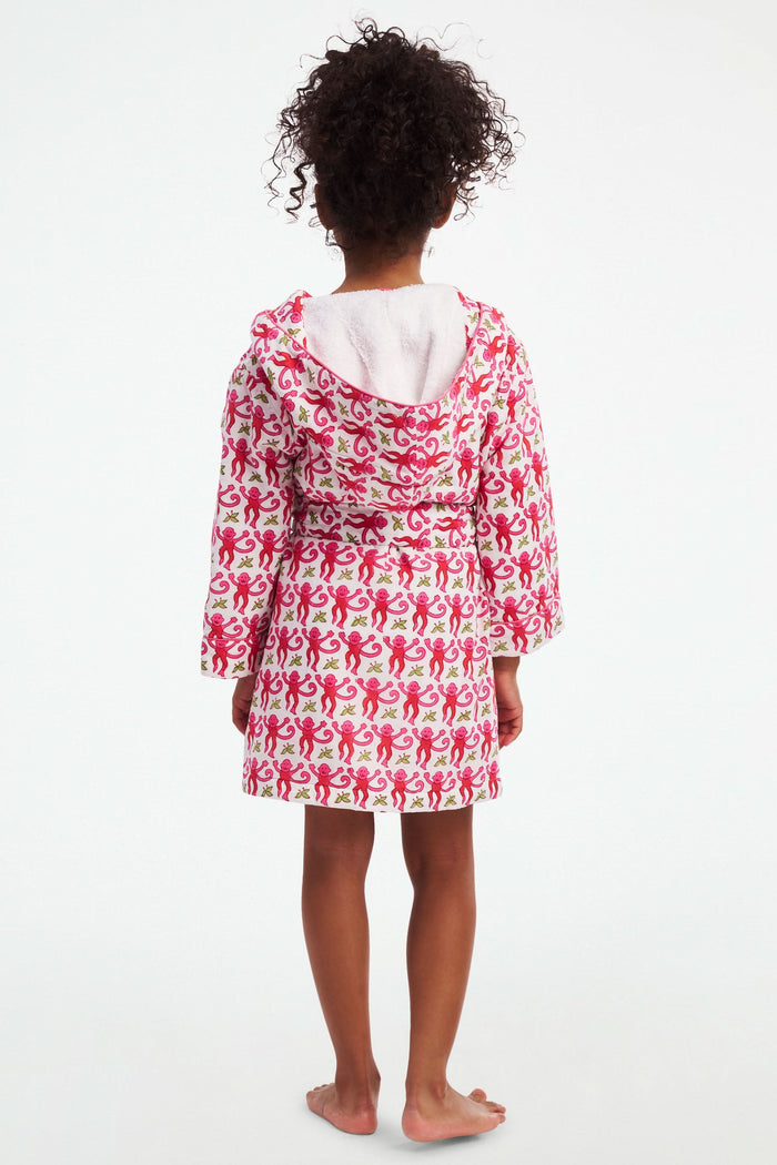 A young girl models the Roller Rabbit Kids Monkey Cosmo Bathrobe, featuring a pink and red monkey print with a hood. She holds the collar with one hand and has her other hand in her pocket, standing against a plain white background.