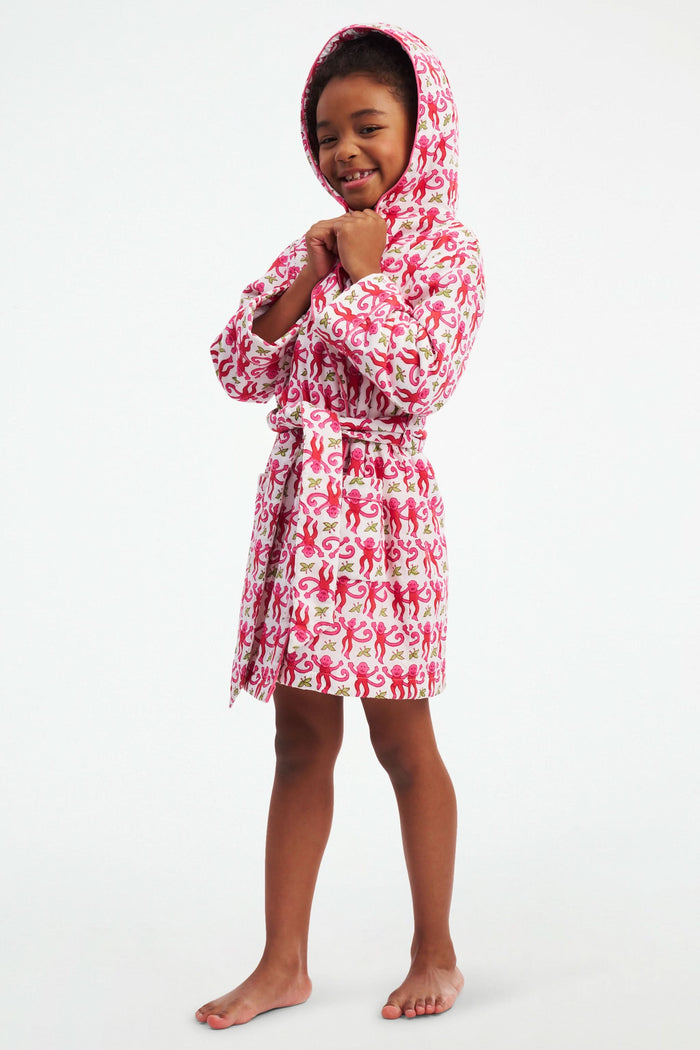 A young girl models the Roller Rabbit Kids Monkey Cosmo Bathrobe, featuring a pink and red monkey print with a hood. She holds the collar with one hand and has her other hand in her pocket, standing against a plain white background.