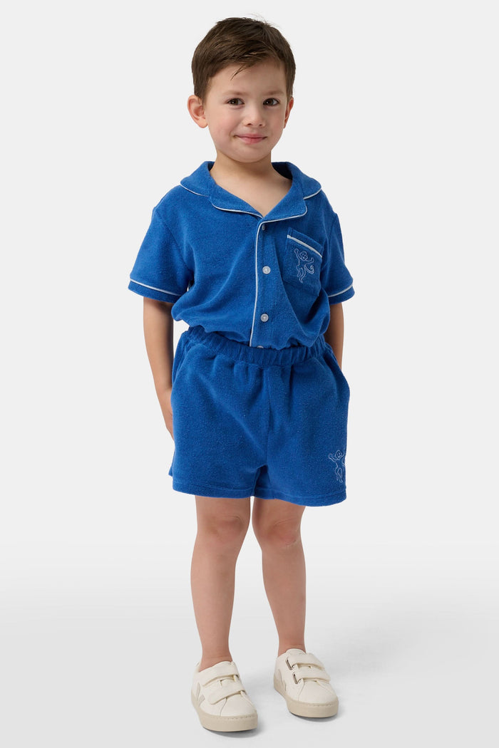 A child wears Roller Rabbit’s Kids Monkey Embroidery Hanni Short in navy with an embroidered figure on the left leg, matching blue top, and white velcro sneakers. Perfect summer shorts for active days.