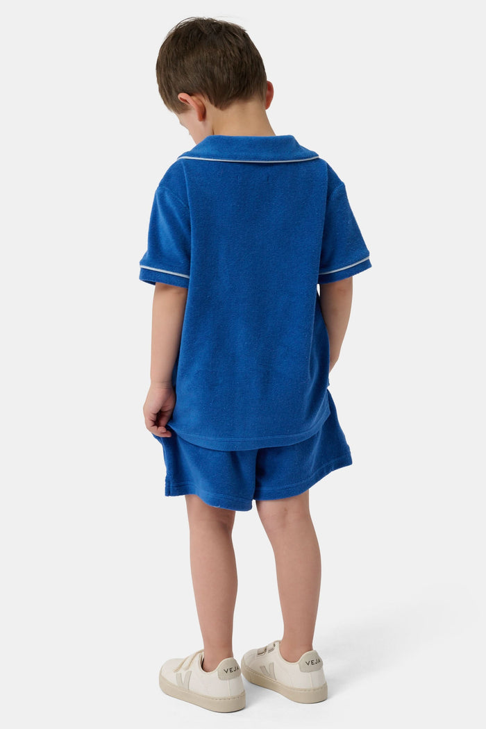 A young child wearing the Roller Rabbit Kids Monkey Embroidery Leo Polo—a navy top with white trim and an embroidered monkey on the chest pocket—smiles with arms raised overhead.