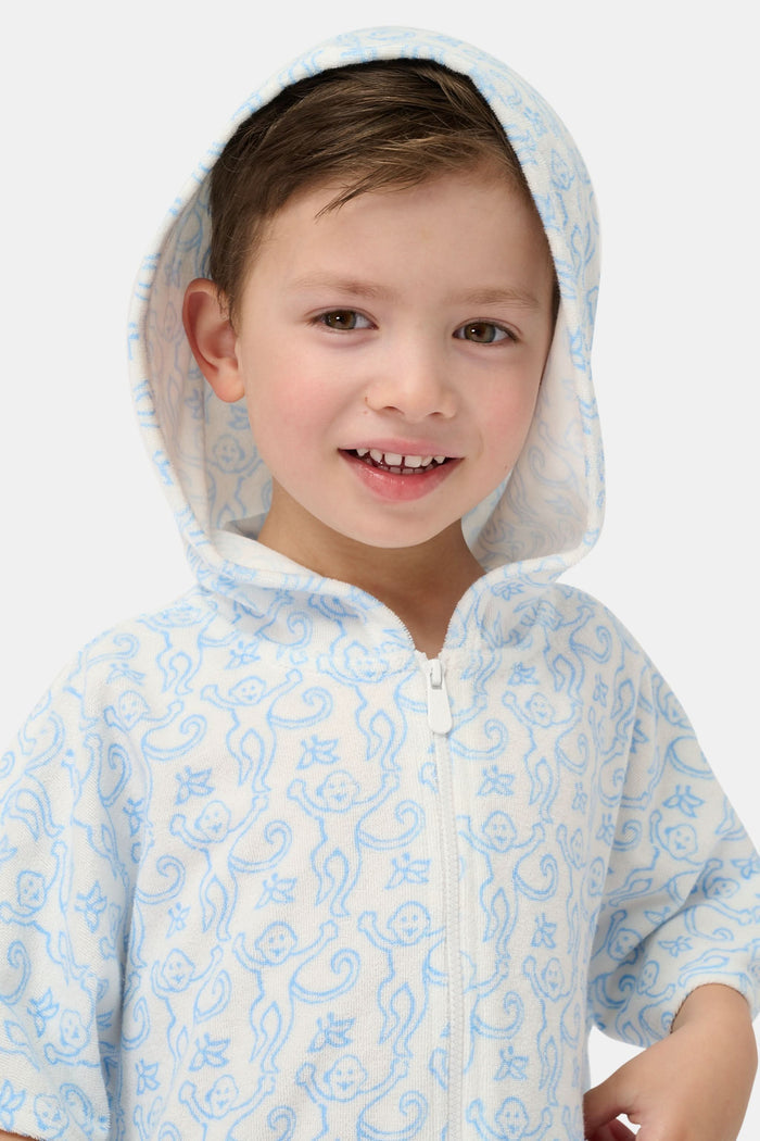 A young child smiles at the camera while wearing the Roller Rabbit Kids Monkey Jesse Cover Up, a white terry zip-up onesie with light blue animal prints and a hood.