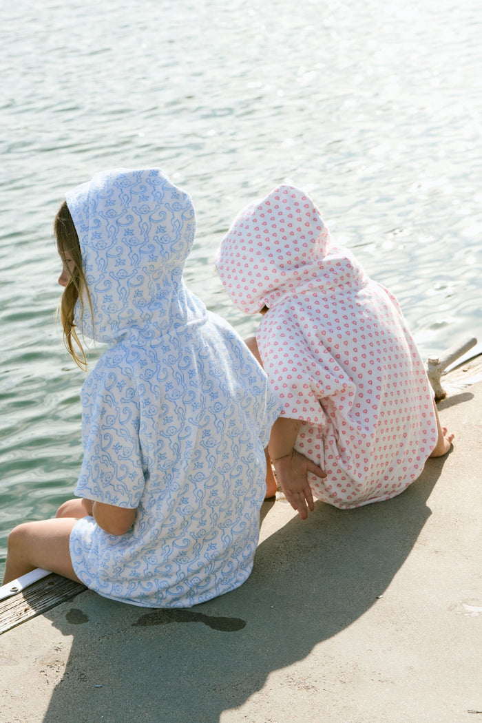 A young child smiles at the camera while wearing the Roller Rabbit Kids Monkey Jesse Cover Up, a white terry zip-up onesie with light blue animal prints and a hood.